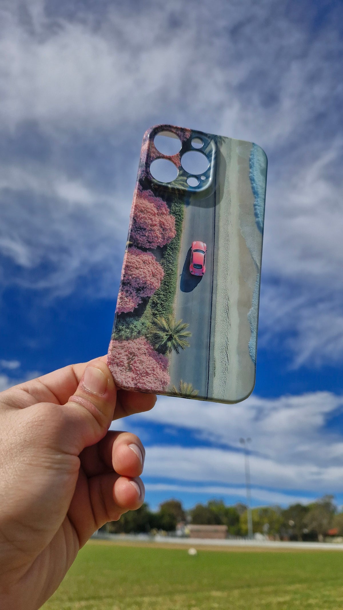 A hand holds a smartphone case outdoors against a bright blue sky with clouds and a grassy field. The case features a printed aerial-style scene with pink trees, a road, and a small red car.